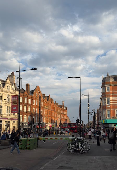 A busy urban street scene on Camden High Street featuring a mix of historical and modern architecture. The foreground shows a paved pedestrian area with several people walking and standing, some near bicycles that are parked and locked to bike stands. The street is lined with low-rise brick buildings having ornate facades, gabled roofs, and large windows, with retail shops and signage visible at ground level. To the right, a multi-storey building with rounded corner architecture and a domed roof is present. Black street lamps and traffic barriers define the edges of the road, which is busy with vehicles including a pink double-decker bus traveling along the street. Large clouds fill the sky, casting diffused natural light over the scene. Waste Removal Camden or similar private waste management services may handle rubbish collected from this commercial and residential area, supporting on-site clearance and alternative waste handling outside of standard municipal collection. Overall, the image depicts a lively area where urban infrastructure and pedestrian activity coexist with the potential for private rubbish removal methods.