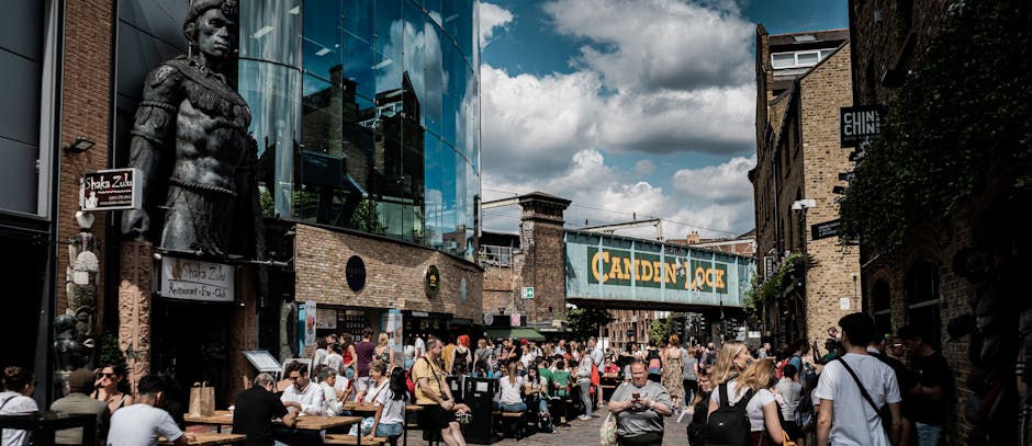 A wide view of Camden High Street showing a busy urban scene with multi-storey buildings lining the street, featuring a mix of traditional brick facades and modern architectural elements. The foreground displays a section of asphalt road with white and red dashed lines indicating traffic lanes and no-parking zones, along with a partially visible pedestrian crossing. On the left side, a scooter is parked near the sidewalk, where a few pedestrians are walking or waiting. The background reveals various small shops and commercial premises with signage, some with retail displays and awnings extending over the pavement. The scene is illuminated by natural daylight with a slightly overcast sky, creating even lighting that emphasizes the textures of the building surfaces and street markings. This environment reflects an active city centre, where independent waste collection by Waste Removal Camden could be part of maintaining cleanliness and managing rubbish from nearby commercial and residential properties, supporting alternate waste handling practices in an urban setting.