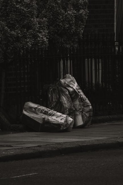 Three large black plastic garbage bags filled with waste sit on the edge of a paved sidewalk, positioned near a black metal fence with vertical bars and a lattice section, beyond which dense foliage of leafy bushes is visible. The bags appear to contain mixed rubbish, with one of the bags partly torn open revealing crumpled materials and plastic packaging. The scene is outdoors, possibly adjacent to a private property or communal area, with the bags placed on a curb or verge next to a road or driveway. The lighting suggests overcast conditions, emphasizing the textured surface of the bags and the surrounding environment. The image relates to private waste management services such as rubbish collection or on-site clearance, as provided by companies like Waste Removal Camden, and reflects typical scenes of out-of-hours rubbish disposal for urban residential settings.